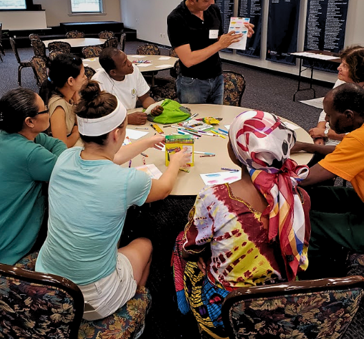 BuildaBridge Institute participants around a table making art while a teaching artist holds up a worksheet